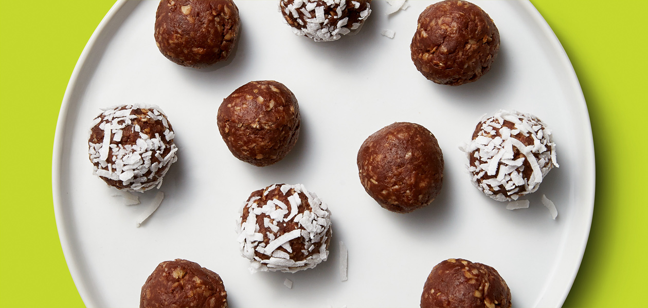 A white plate contains a pattern of 10 small banana protein balls, half coated with vibrant white coconut flakes and half plain and chocolatey.