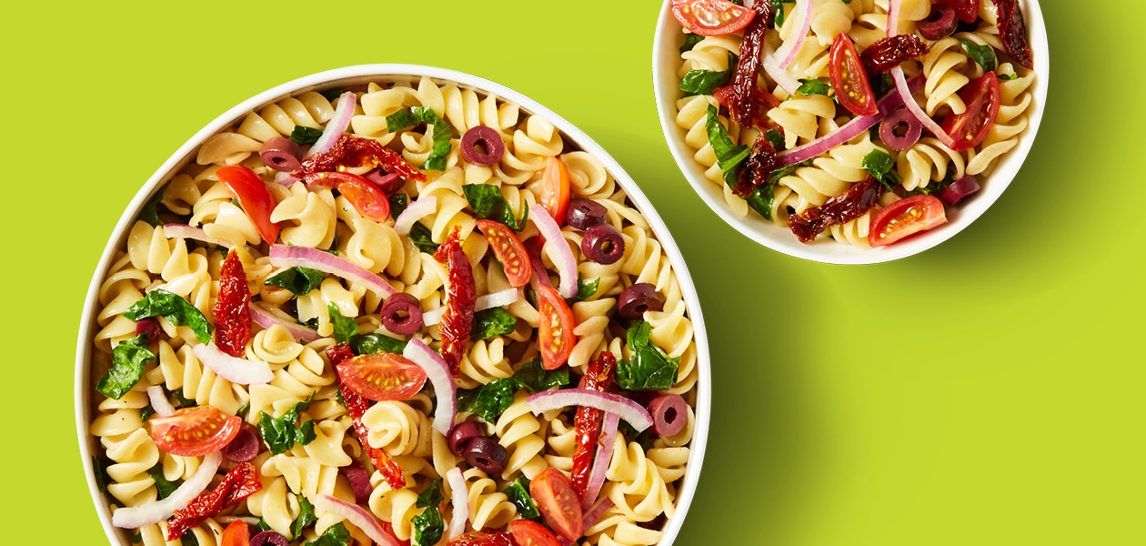 Two pasta dishes, each containing a generous portion of pasta salad with fussili, tomatoes, onions, spinach, and olives deliciously mixed together.